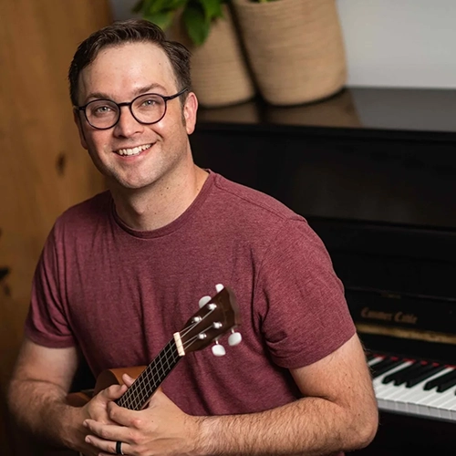Matt Hobbs holding a ukulele, piano in the background. 