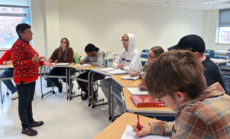 Faculty member lecturing to a group of students sitting at desks in a semicircle.