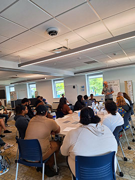 Students sitting at tables, listening to a professor positioned at the front of the room.