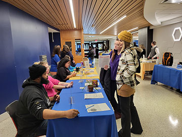 Students at Orientation talking to College administators who are sitting at tables in the Begley Building.