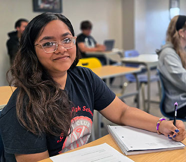 A female ECSH student with long dark hair and wearing glasses, seated at a desk, writing in a notebook.