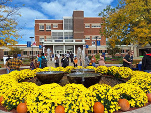 Quad fountain surrounded by yellow mums and pumpkins, lots of students milling around in the background. 