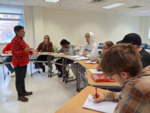 Faculty member with students seated at desks in a semicircle around her. 