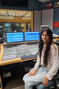 Female student seated at the soundboard in the recording studio, student on a piano in the background.