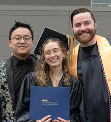 Three graduates pose in the caps and gowns, the grad in the center holding a diploma.