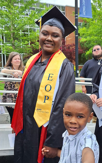 EOP graduate in cap, gown and EOP stoll stands at commencement holding her young son's hand.