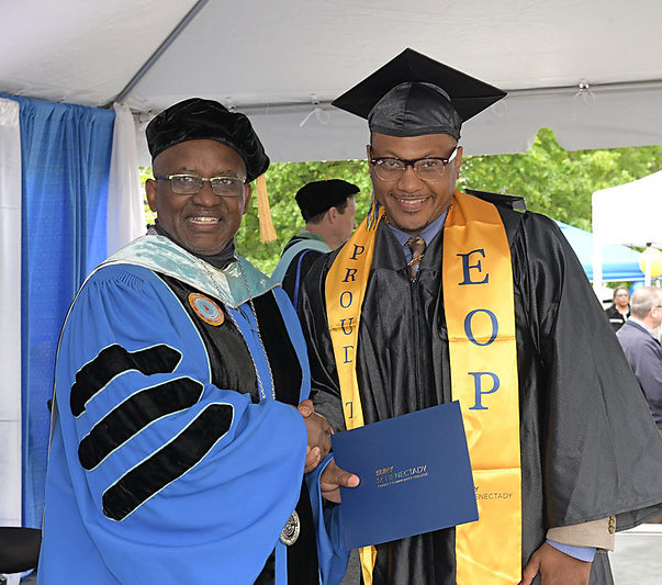 SUNY Schenectady President, Dr. Steady Moono, stands with a graduate in a cap and gown and an EOP stoll.
