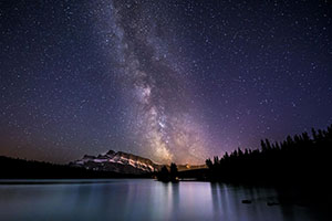 Night sky filled with stars, a lake reflects the sky in the foreground.