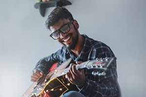 Man wearing glasses, playing the guitar with a smile on his face..