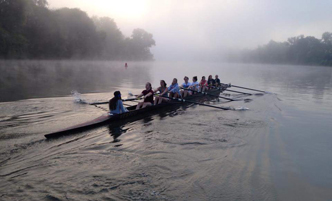 Women's crew in an 8-person boat on a misty river.