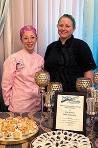 Chefs Rain O'Donnell and Vanessa Schulte stand behind a table filled with individual desserts. 