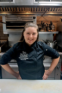 Chef Cassondra Saddlemire, wearing a dark chef coat, stands with hands on hips in a restaurant kitchen.