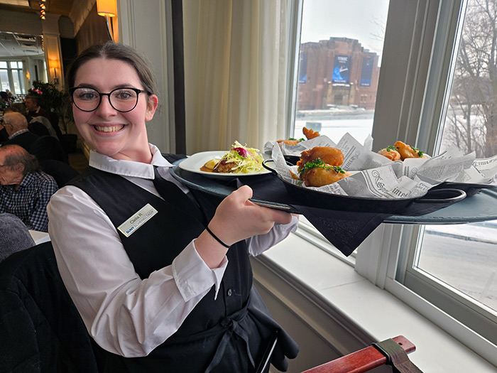 Hospitality student in a white shirt and black vest, holding a tray of plated meals inside the Casola Dining Room.
