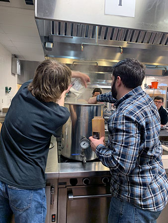 Two students working over a brew pot that is steaming. 