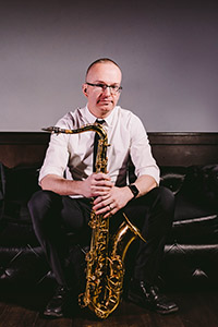 Brian Patneaude sitting on a leather couch, holding a saxophone.