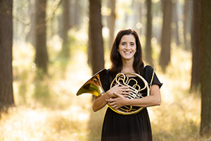 Catherine Pandori holding a horn, standing in forest.