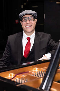David Gleason sitting at a piano, wearing a black suit, red tie, and a newsboy cap.