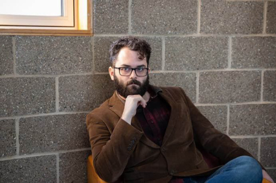 John Carroll in a brown jacket, sitting in a chair. 