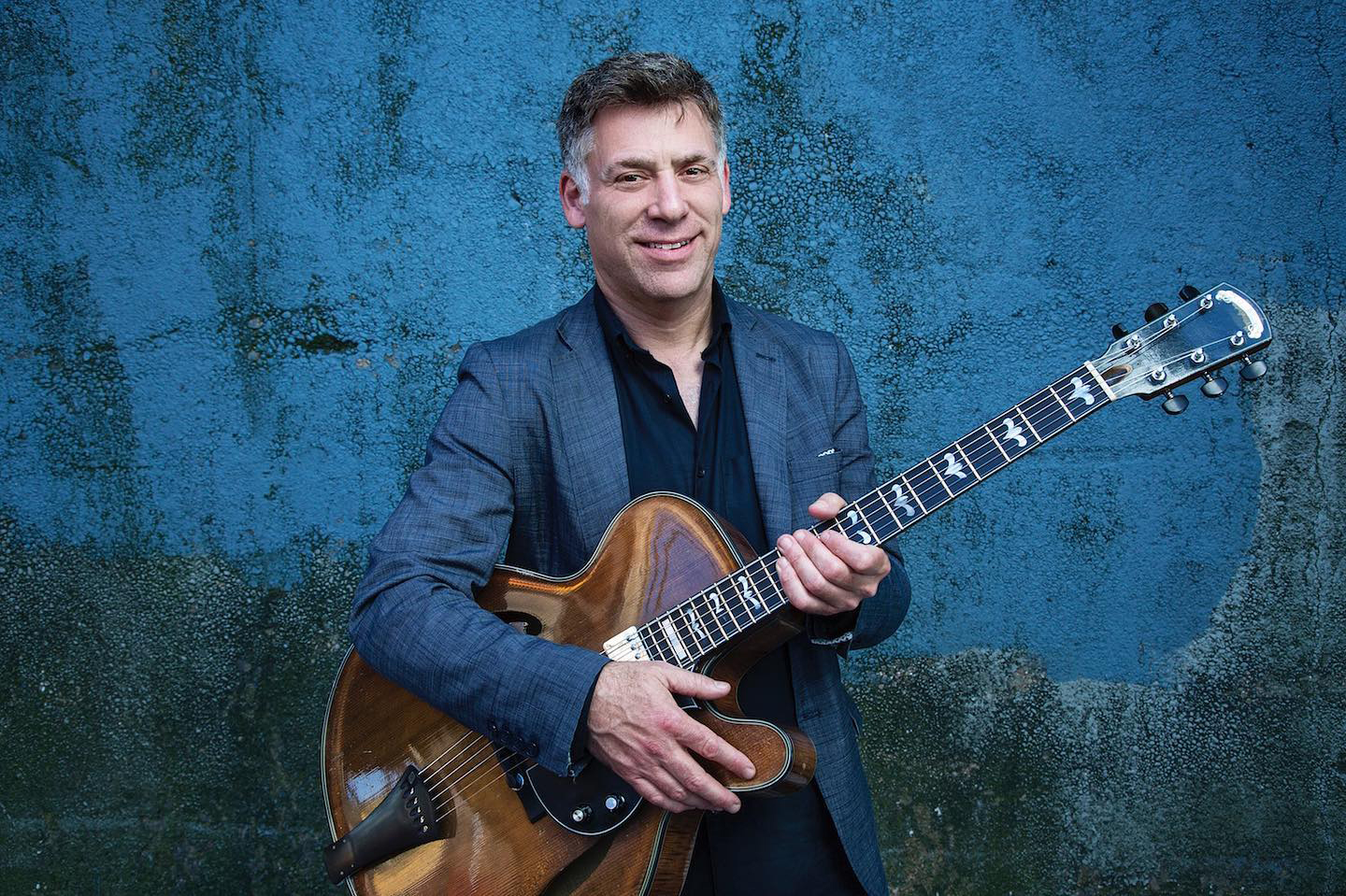 Peter Bernstein in a black shirt and dark blue jacket, holding a guitar. 