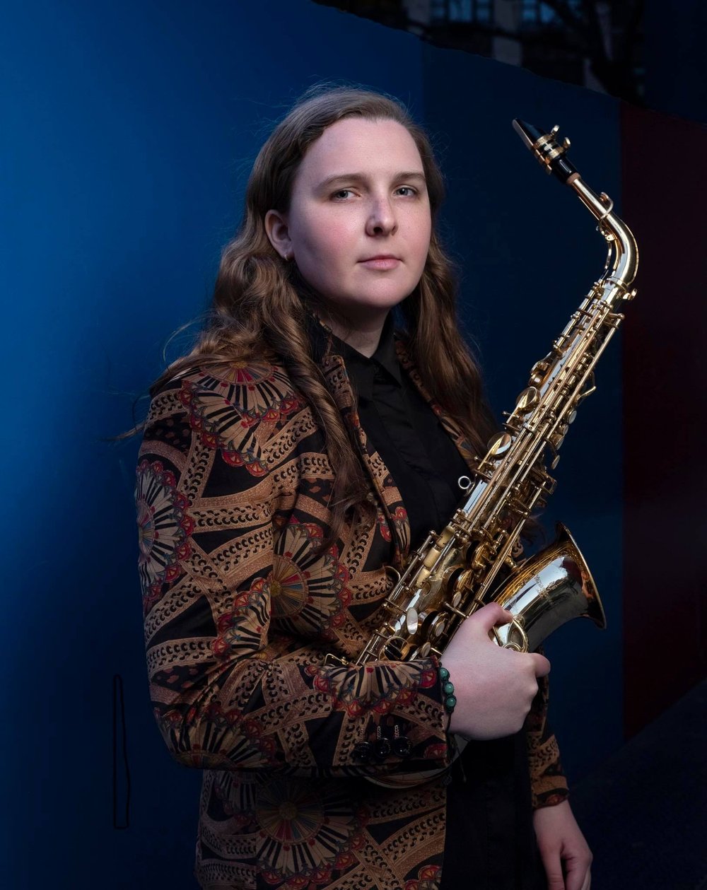 Sarah Hanahan in a brown patterned suit coat, holding a saxophone.