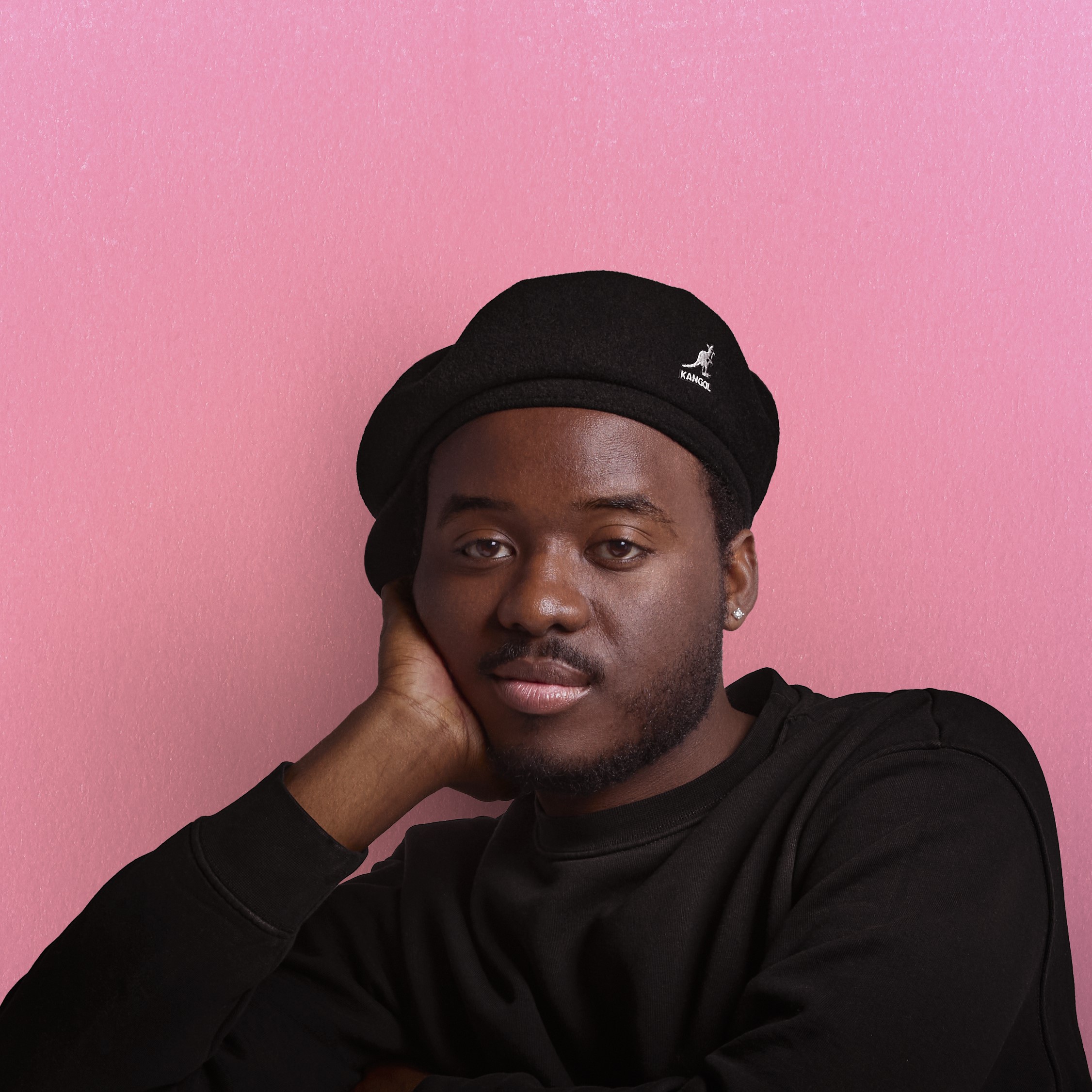 Tyreek McDole in a black shirt and black beret, leaning against a pink wall.