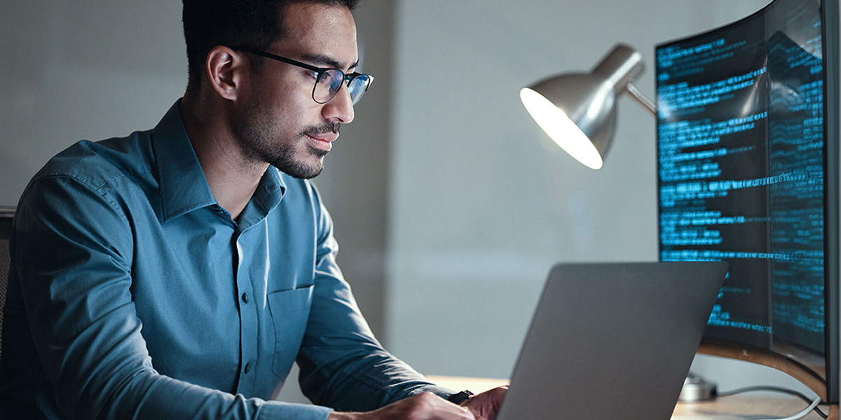 Man working on a laptop with large curved monitor attached showing lines of code.