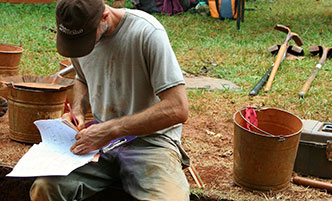 Archaeology student sitting on the edge of a dig pit, doing paperwork.
