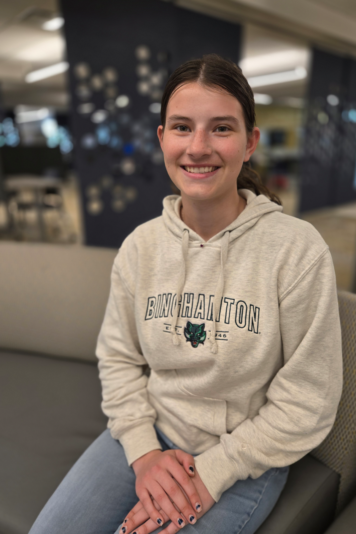 Grace Van Baaren in a Binghamton University sweatshirt, sitting in the Begley Learning Commons..