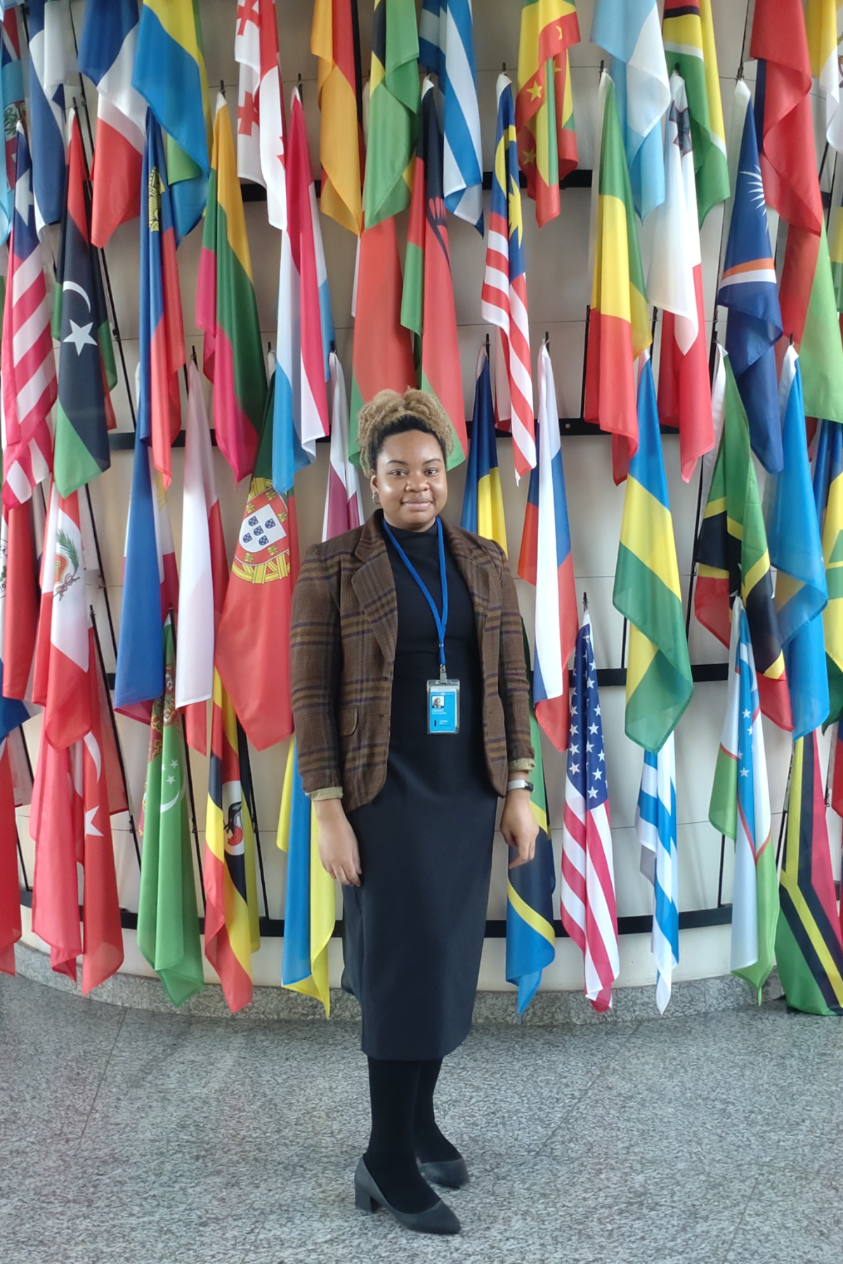 Nkeiru Ubadike standing, smiling with international flags behind her, inside the International Atomic Energy Agency Headquarters in Vienna, Austria, where she is interning.