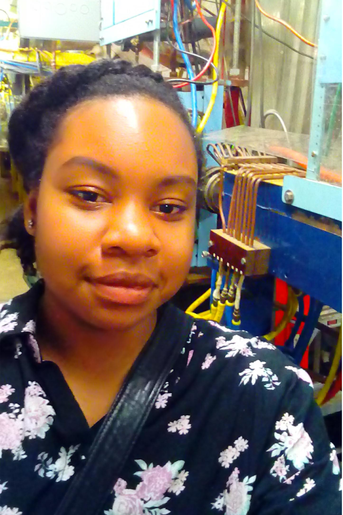 Nkeiru Ubadike standing facing camera, in front of a particle accelerator component at Cornell Electron Storage Ring at Cornell University in 2018.