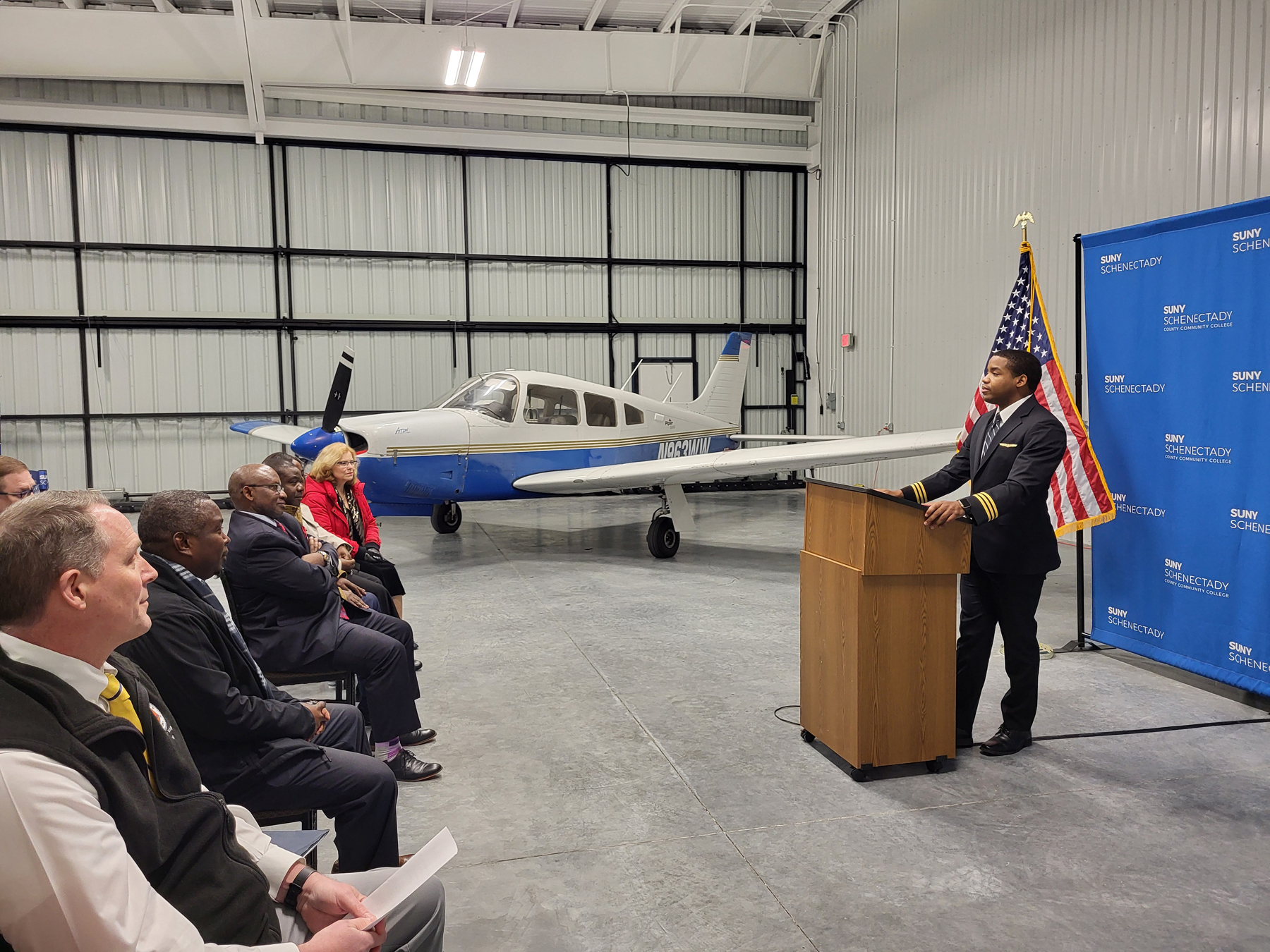 A pilot stands at a lecturn speaking to an audience in the new SUNY Schenectady hanger. A small airplane is parked in the background.