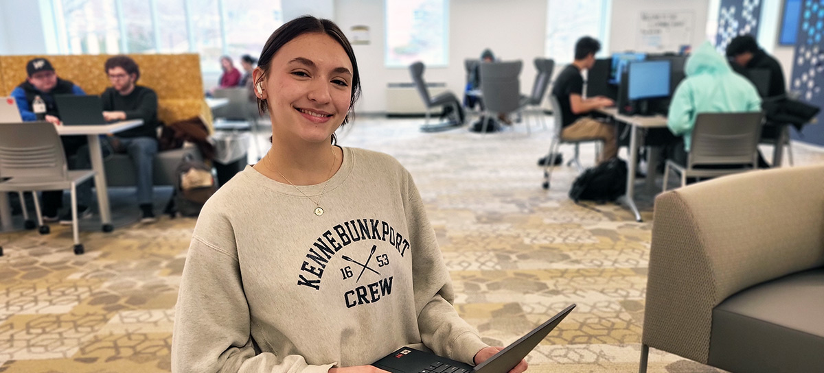 Female student sitting with a laptop in her lap, other students working at tables in the background.